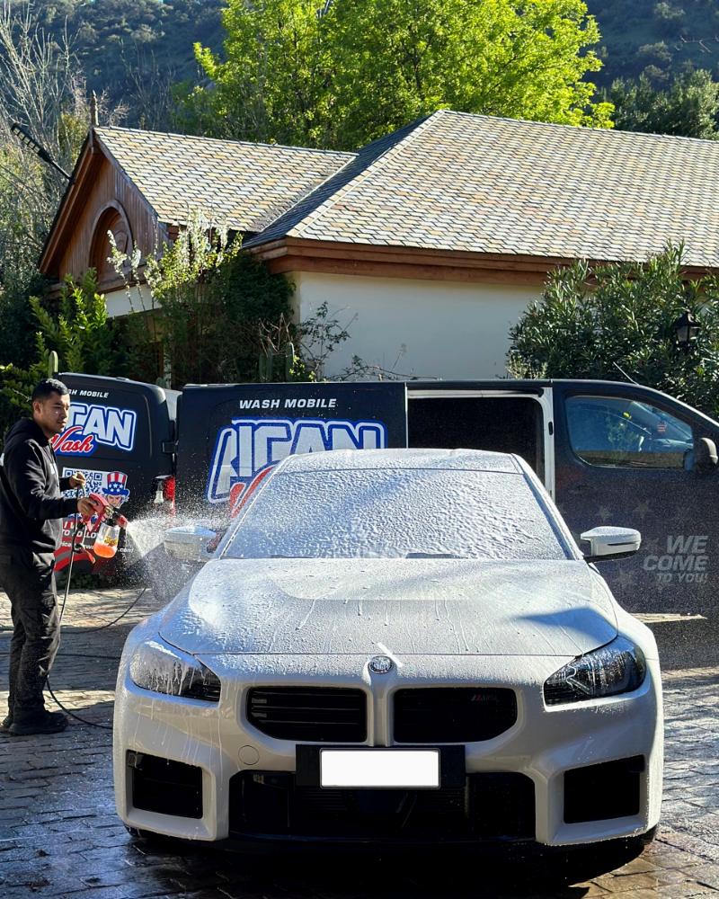 Un BMW cubierto de espuma frente a una casa; detrás, la van de American Car Wash con la puerta abierta y una persona del equipo aplicando espuma